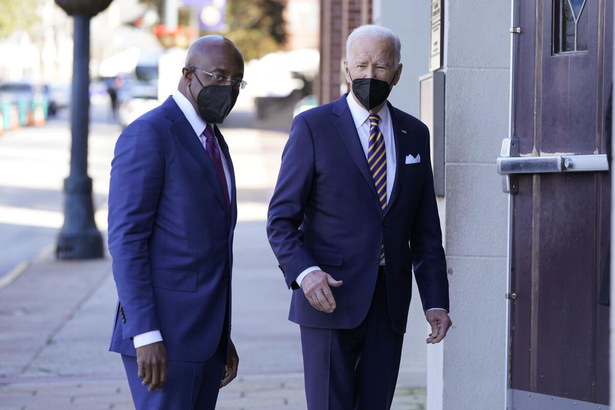 Sen. Raphael Warnock and President Biden approaching a building entry from a city sidewalk 