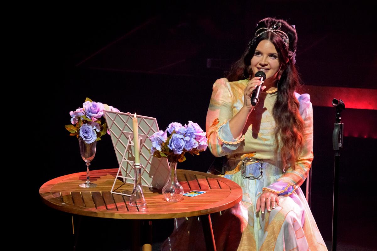 A woman seated at a cafe table that holds vases of flowers and a candle sings into a handheld microphone