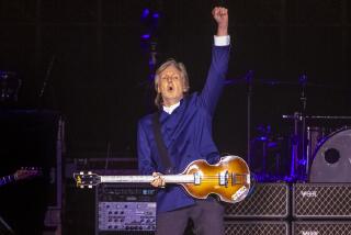 Paul McCartney in a blue long-sleeve shirt on a stage holding up his left hand in a fist, while holding a guitar