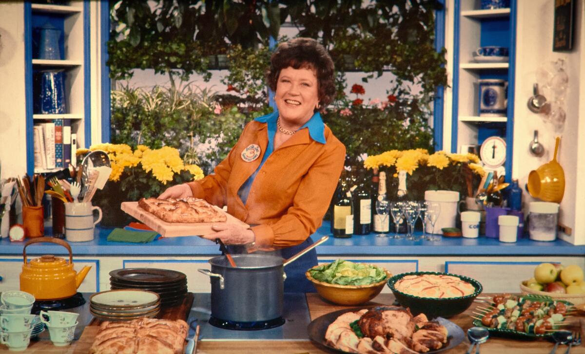 Julia Child stands in a kitchen surrounded by various prepared dishes and bottles of wine..