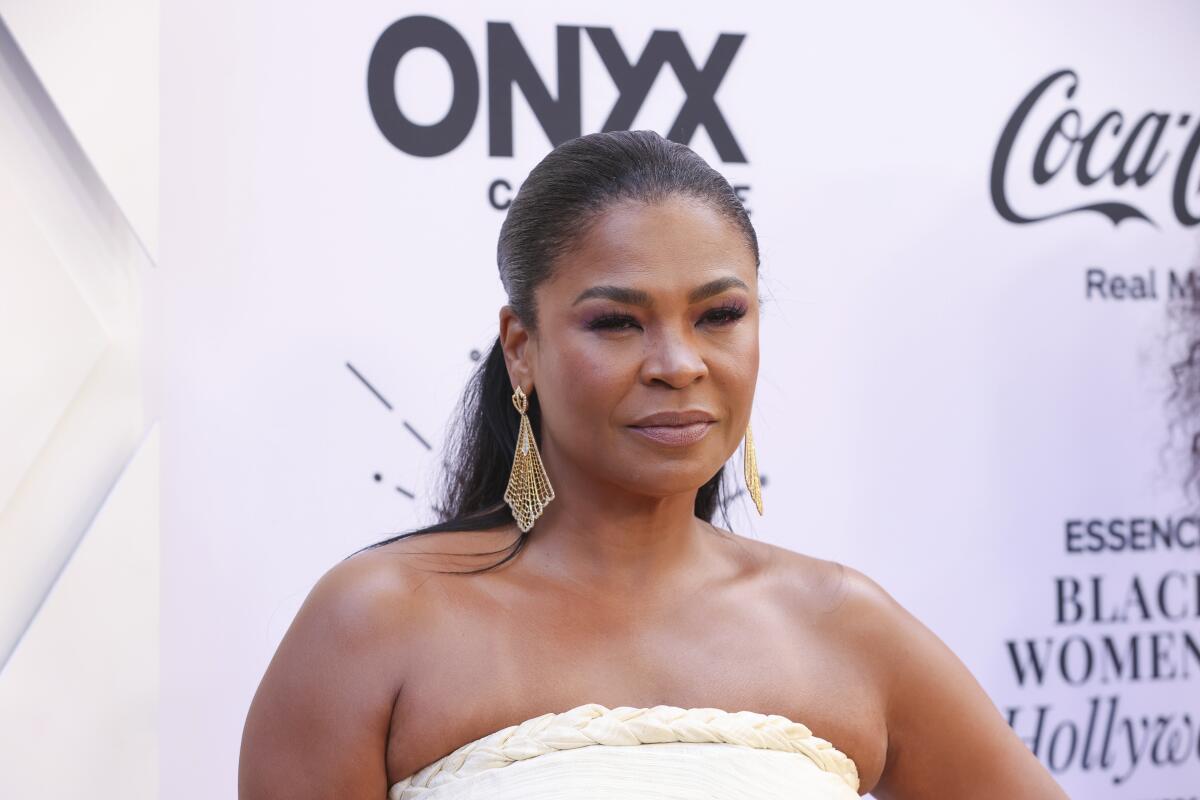 A woman wearing a ponytail, a strapless top and chandelier earrings poses in front of a backdrop