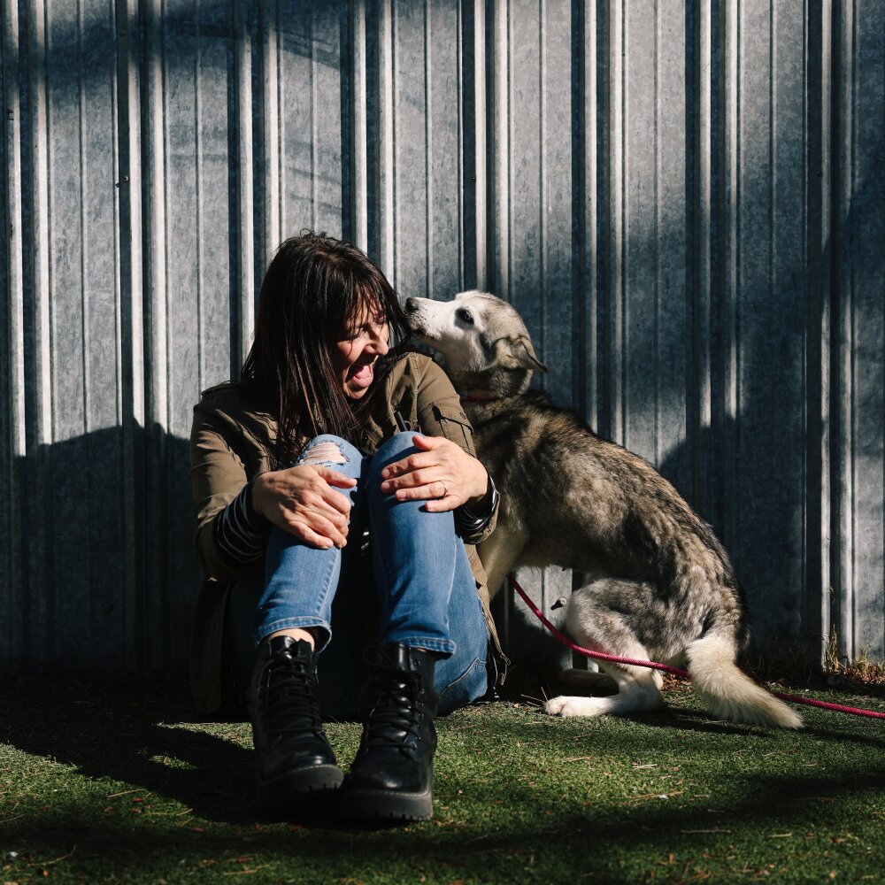 Rita Earl Blackwell sits on grass and smiles as a large gray dog sniffs her.