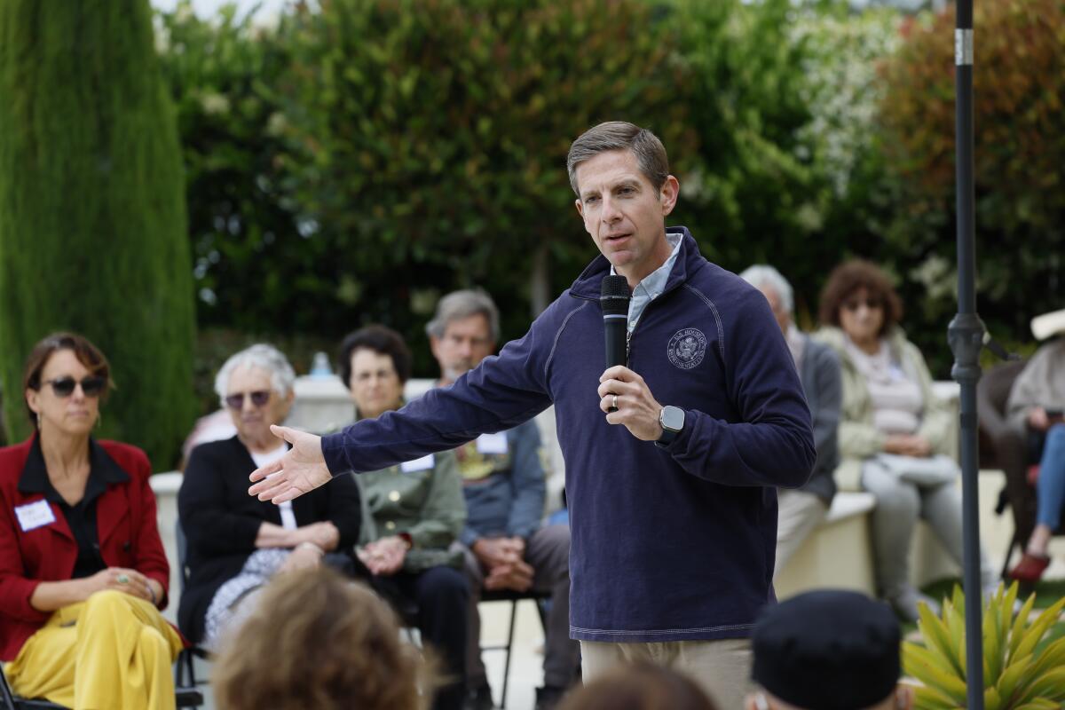 Mike Levin stands outside, one arm out, speaking into a mic to a small crowd seated around him against green trees and shrubs