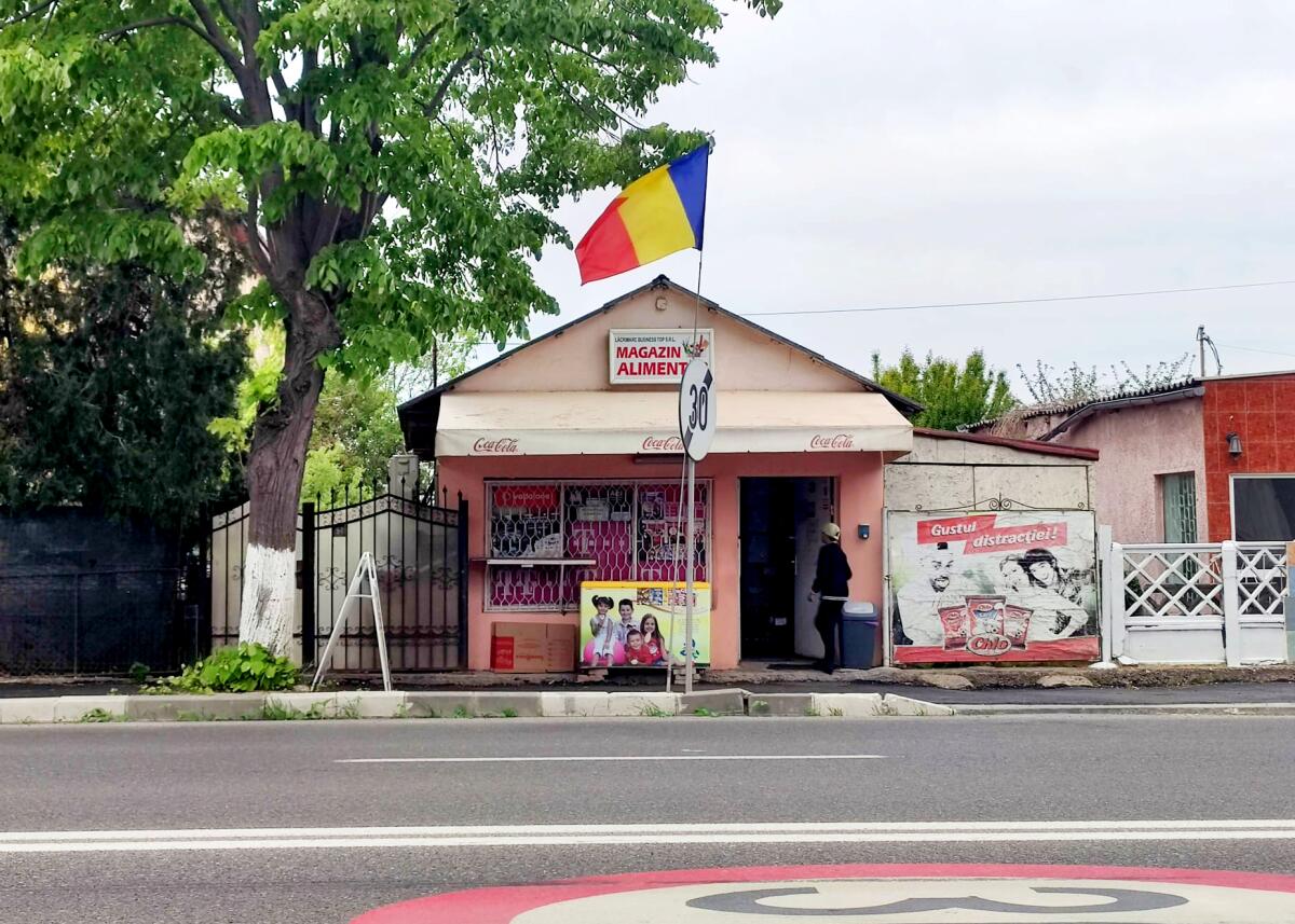 An exterior of a small building painted pink, with the Romanian flag perched above its entrance. A person is entering.