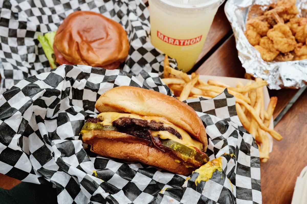 A smash burger at Marathon Burger. Behind is a Marathon-branded cup of lemonade and a basket of fries, and fried shrimp