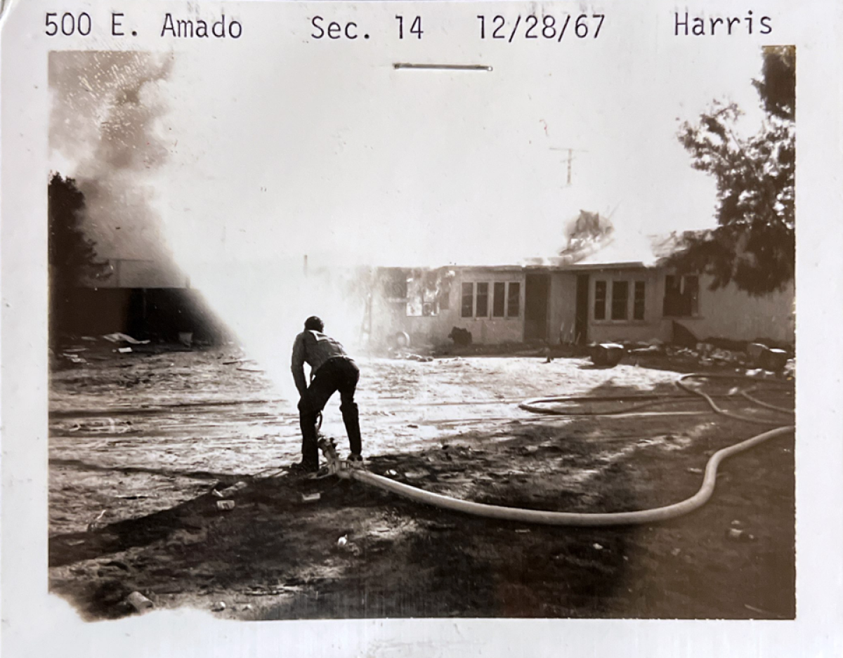 A black and white photo of a house on fire as someone sprays a firehose in the foreground.
