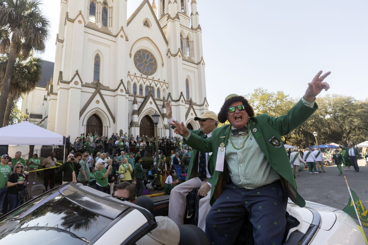 A person in a convertible rides in a parade route along a crowd wearing green