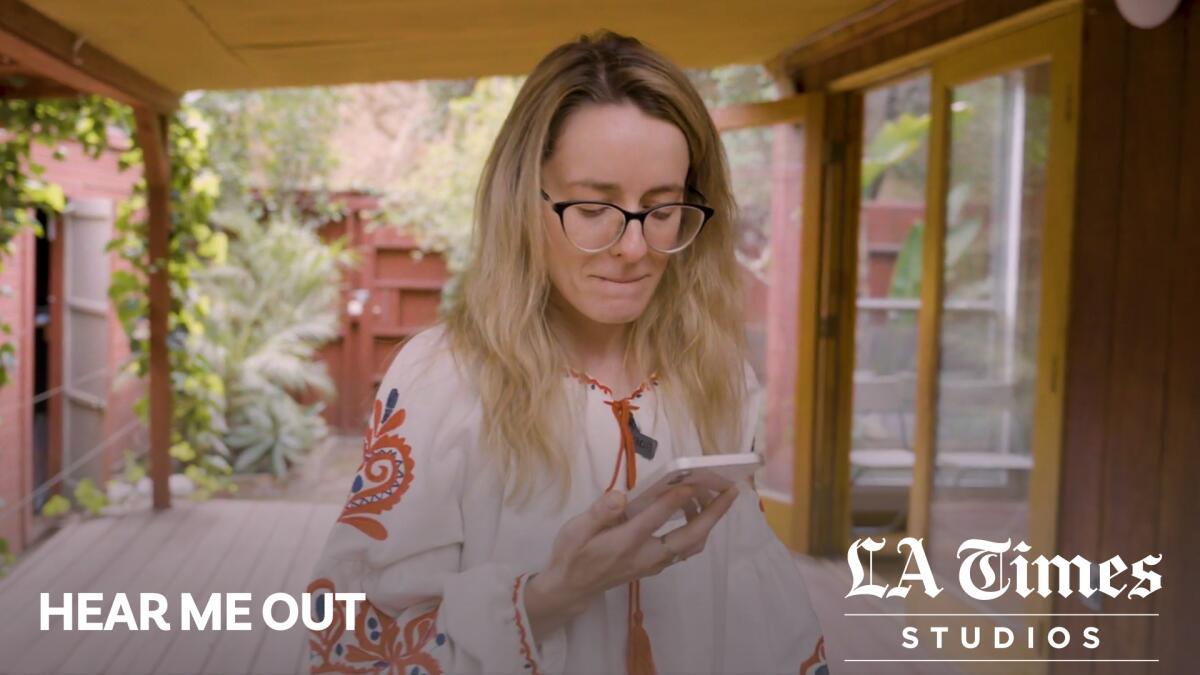 A blond woman wearing glasses looks at a phone screen in a backyard