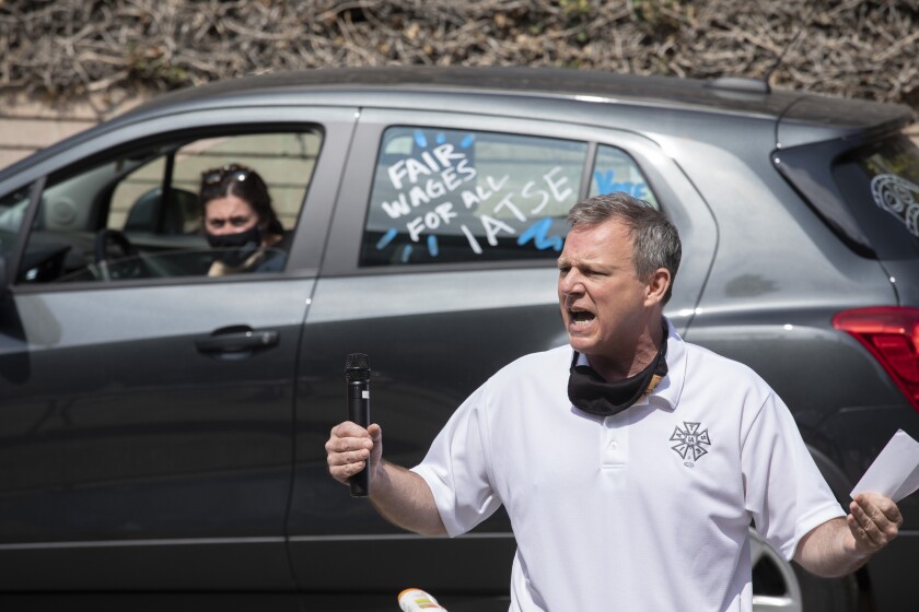 A man yells out. A car with "Fair wages for all IATSE" is in the background