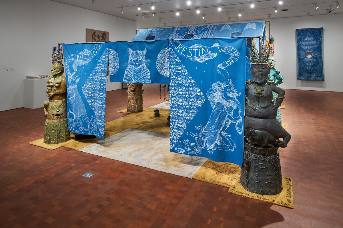 An indigo-colored tent bearing images of cats and flanked by ceramic figures occupies a central space in a gallery.