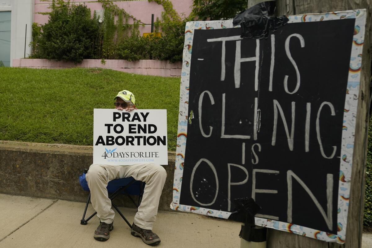 An abortion opponent sits in a chair holding a protest sign outside a clinic with a sign reading "This clinic is open"