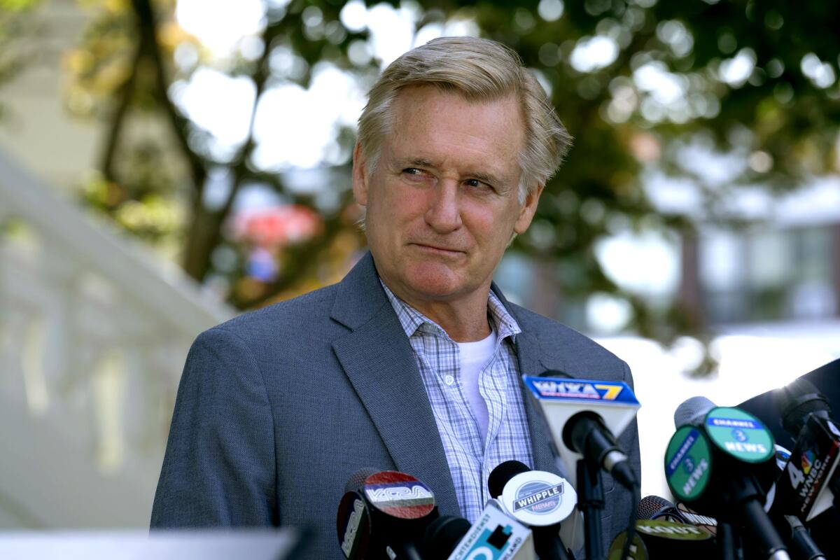 A man in a blue blazer and plaid shirt stands before microphones at a press conference.