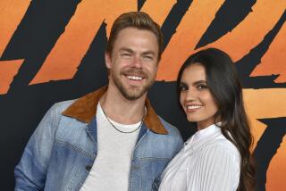 Derek Hough dressed in denim jacket poses next to Hayley Erbert in a white dress as the pair embrace and smile
