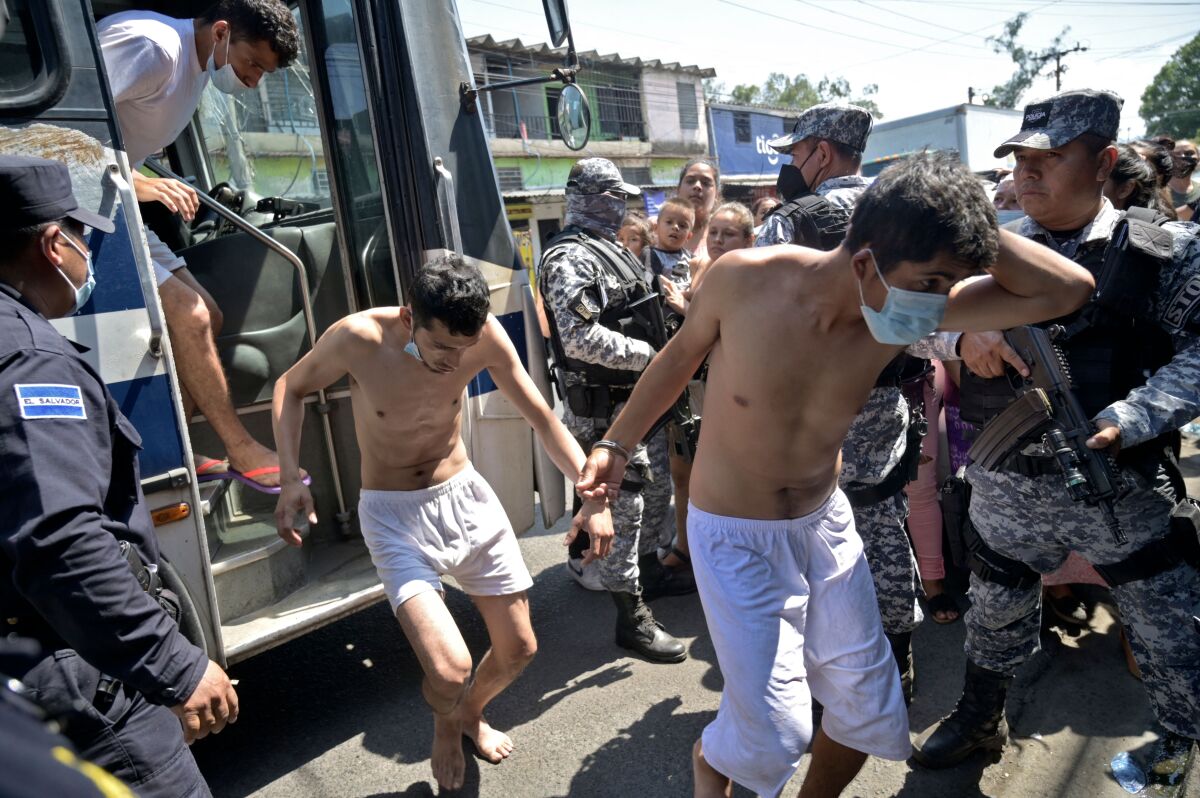 Men in white shorts and no shirt disembark from a bus with armed men in fatigues standing watch.
