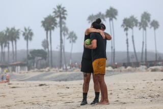 Two people, one holding a small watermelon, embrace on a beach, with palm trees behind them