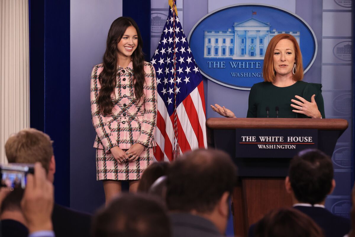 A woman in a dark dress, right, standing at a White House lectern, gestures next to a smiling young woman with dark hair