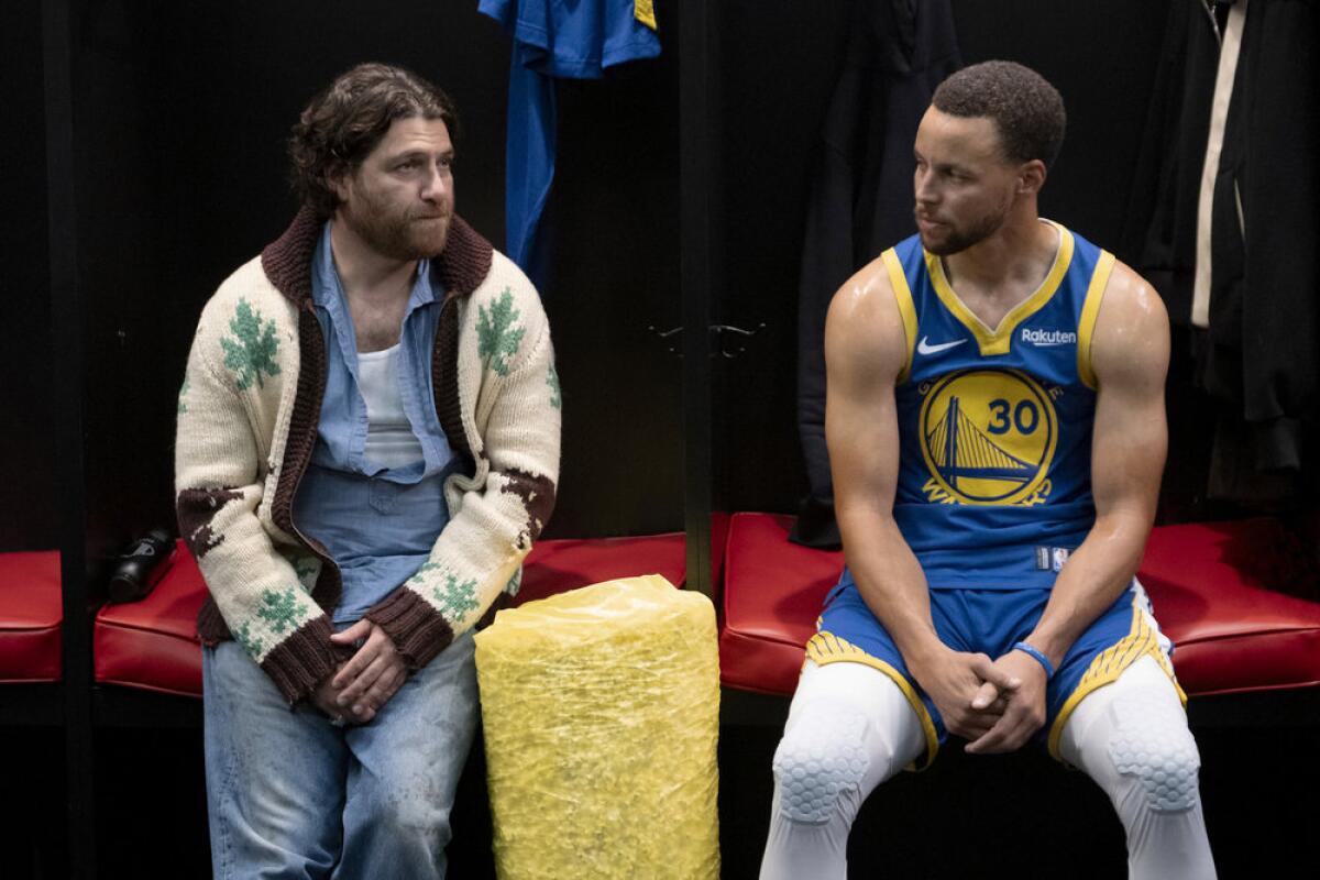 A man in a white and brown sweater sitting next to Steph Curry in a blue and yellow basketball uniform.