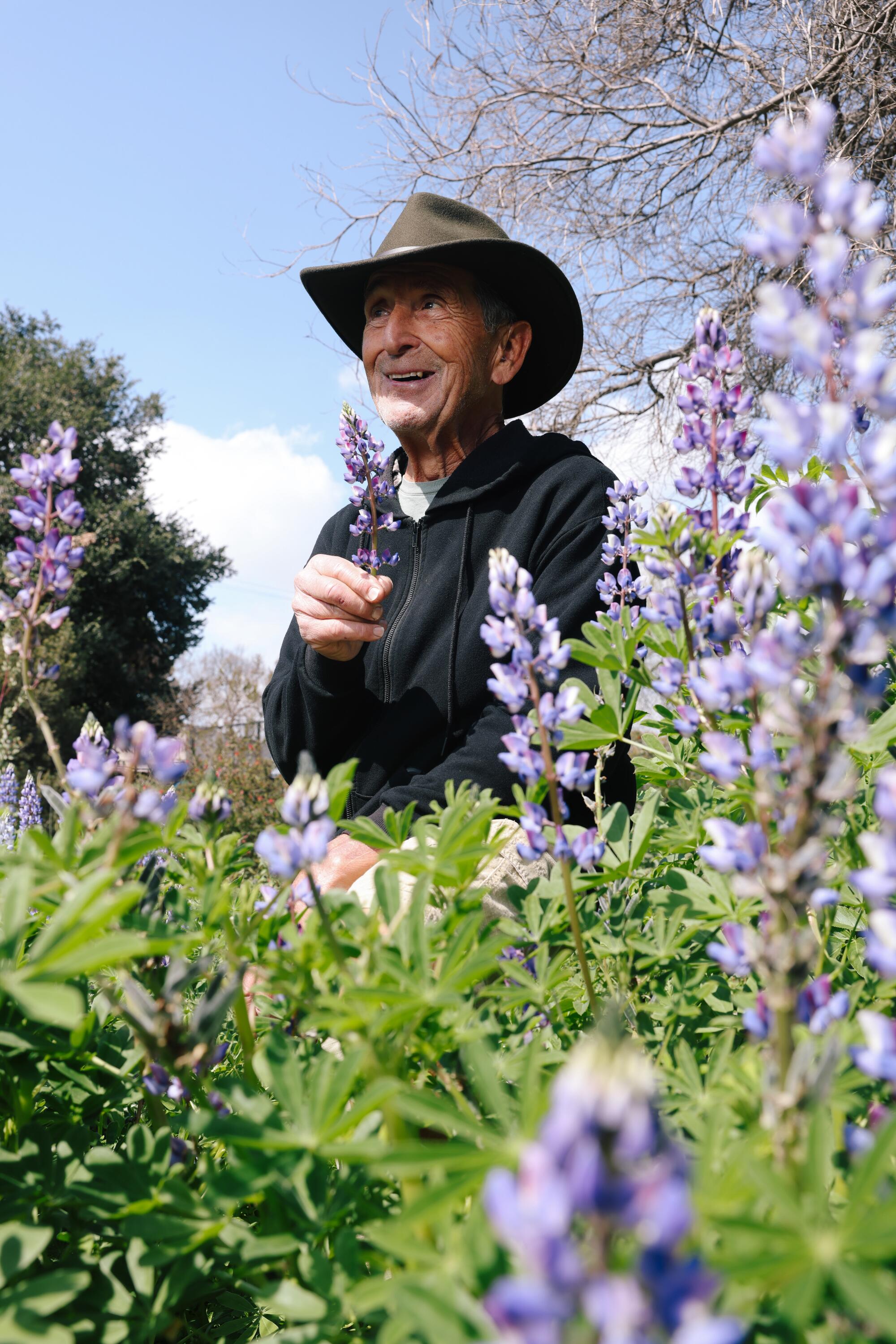 A man standing in a garden smiling and holding a flower in  his hand