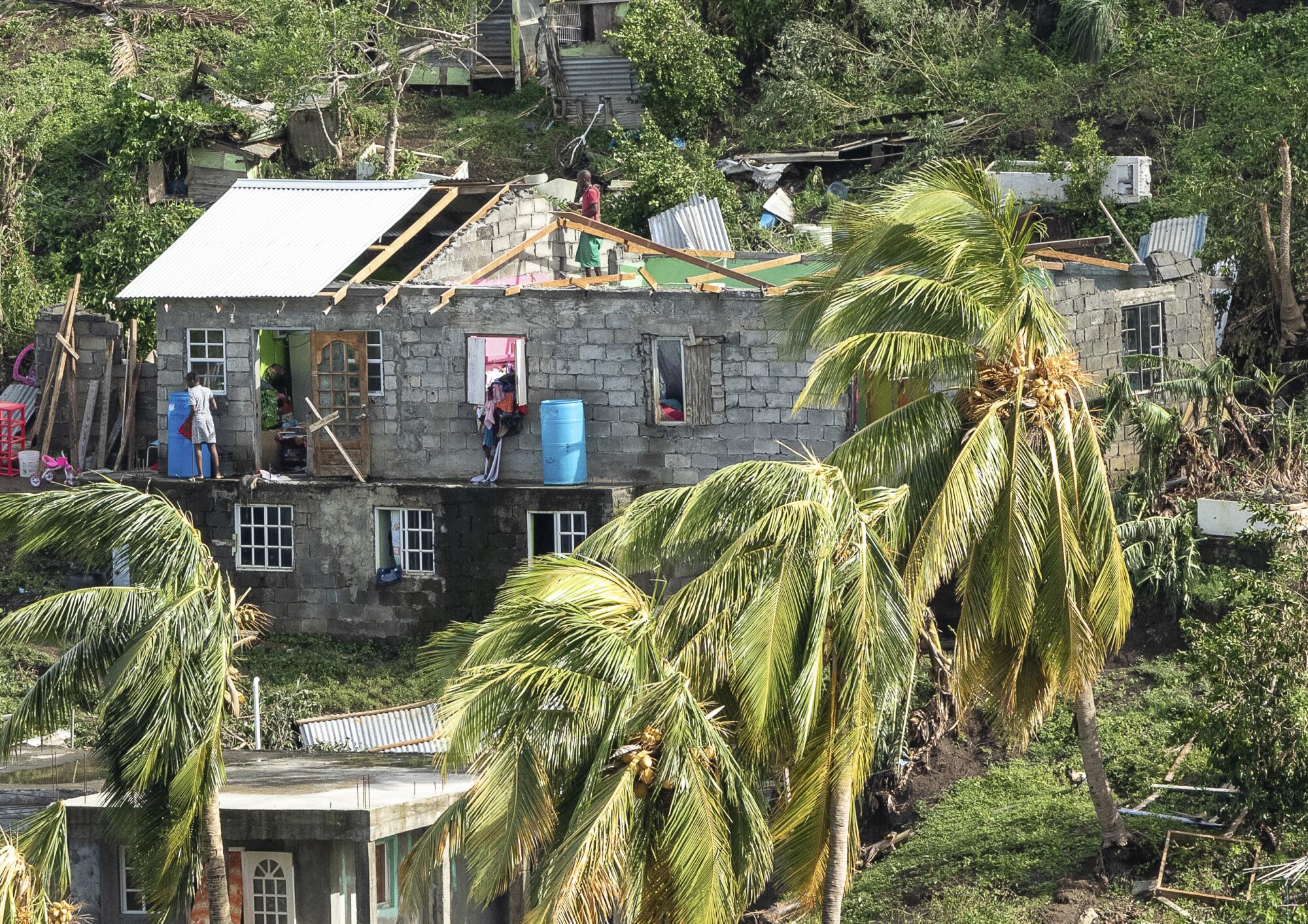 People working on the upper level of a home with a damaged roof, with palm trees blowing
