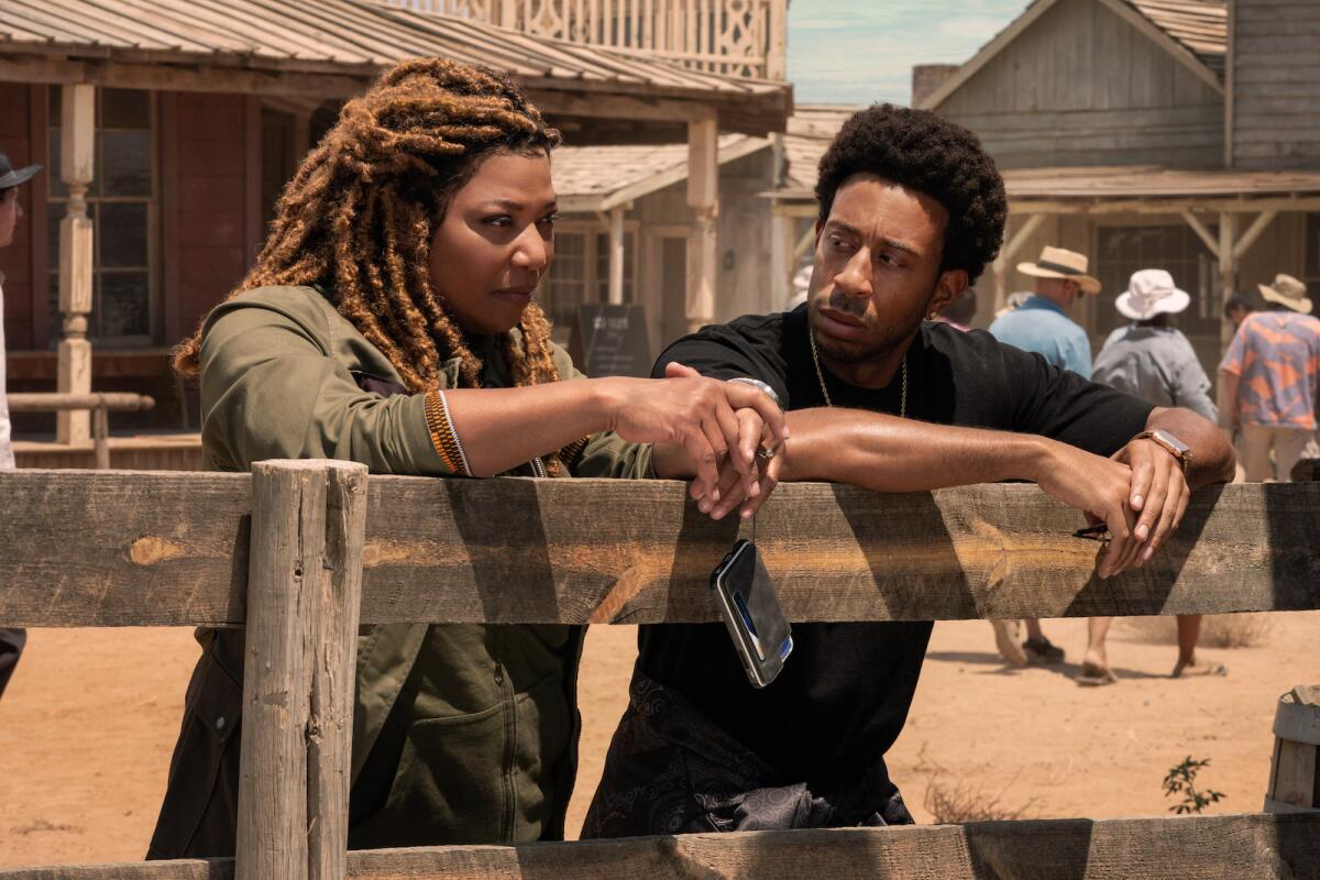 A man and woman having a serious conversation lean against a fence in a dusty Western town. 