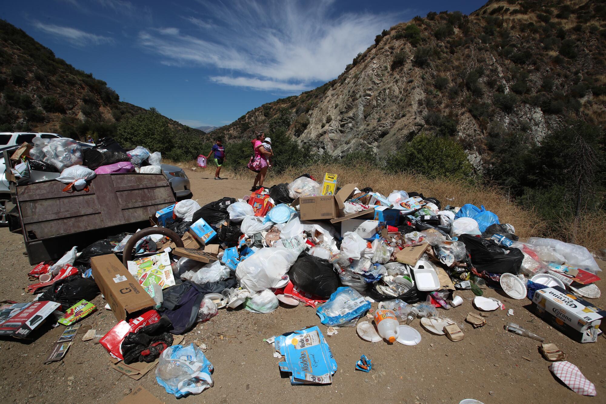 Heaps of trash are piled near a garbage bin as mountains rise in the background.