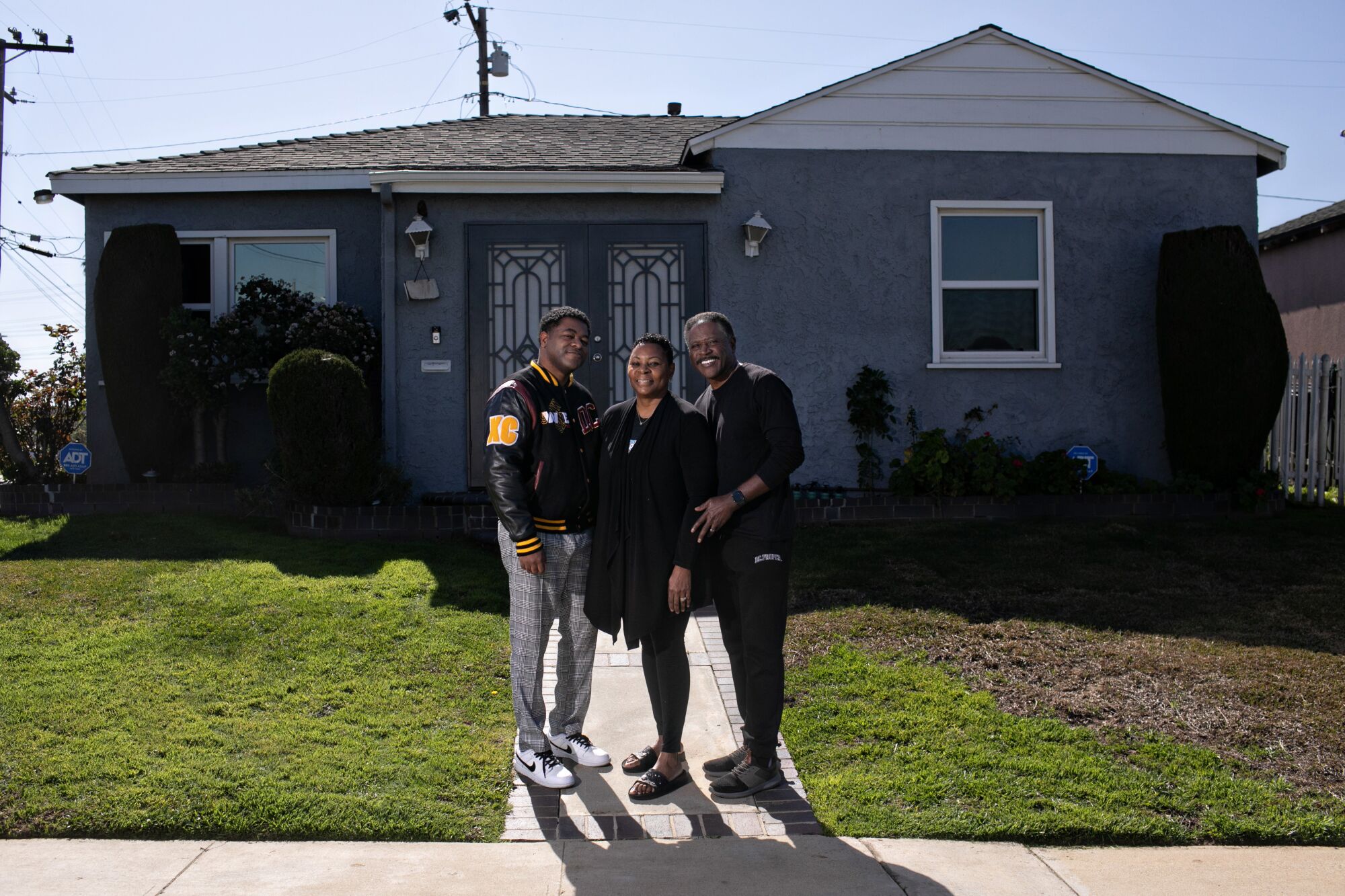 A man stands with his parents outside a modest house.