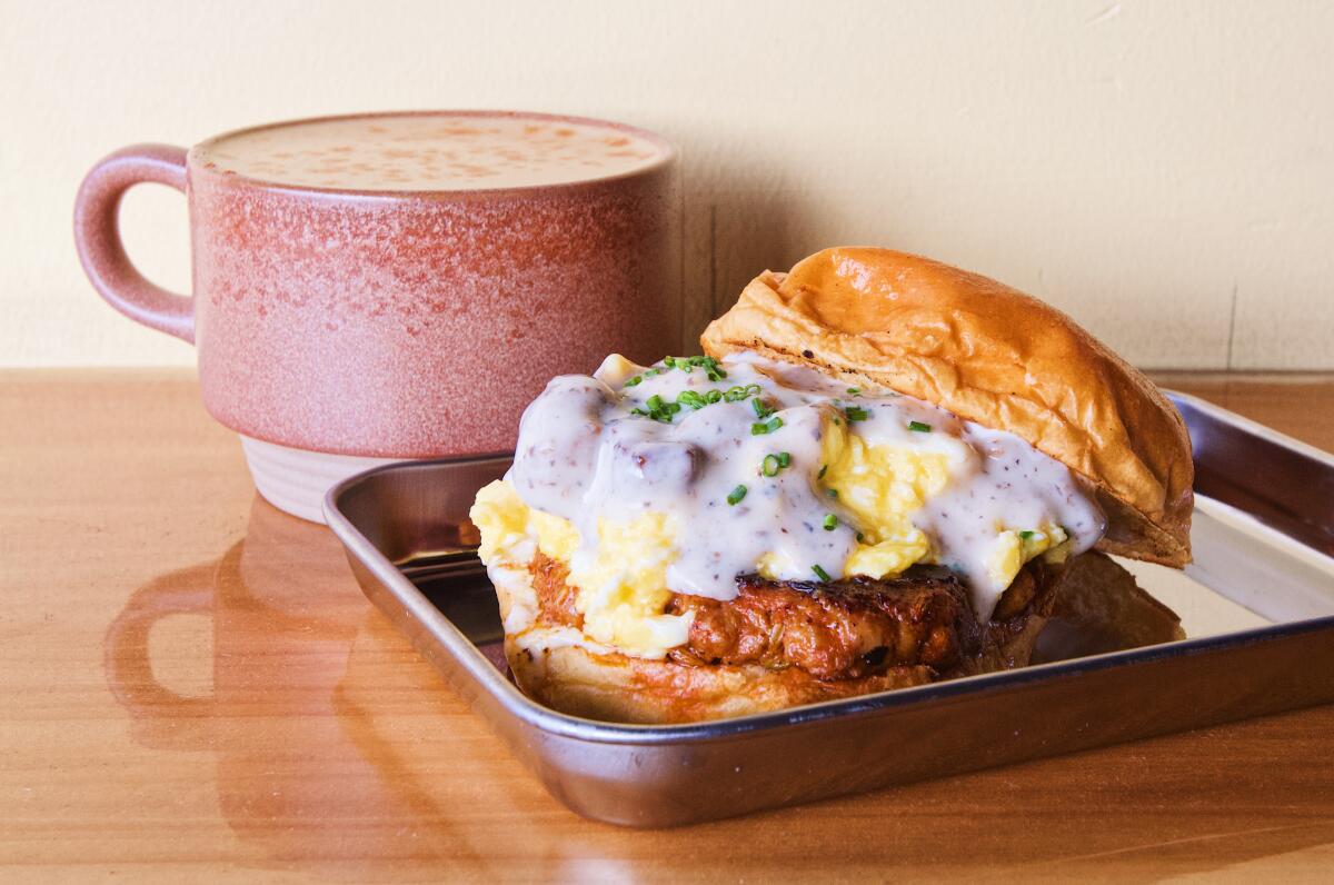 A photo of a chorizo breakfast sandwich topped with  eggs and gravy. In the background is a pink ceramic mug of cafe de olla.