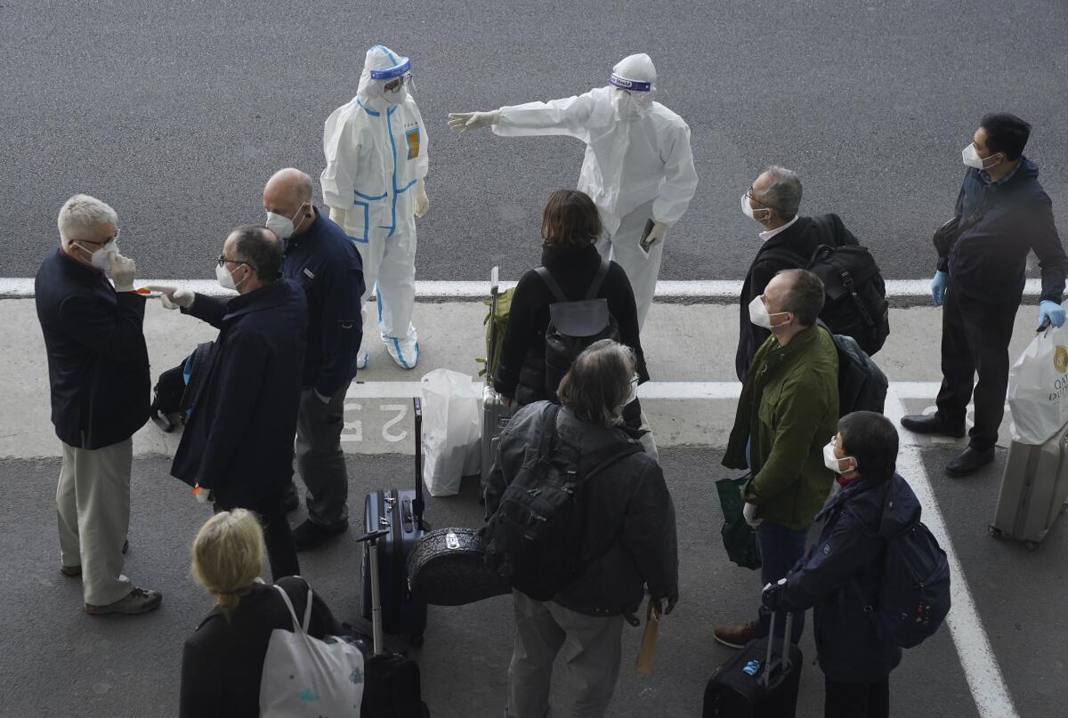 A group of people with suitcases stand in front of two people in head-to-toe protective gear.