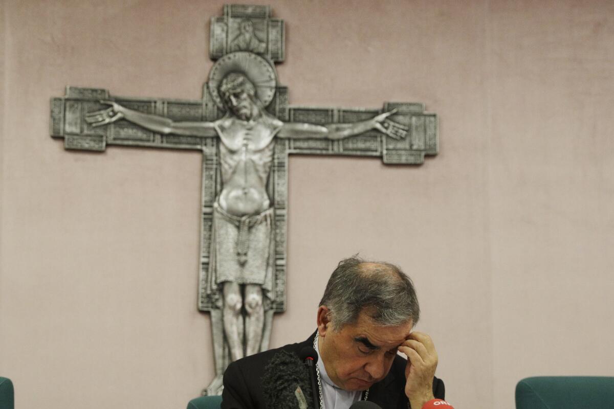 A cardinal sits below a large crucifix with his head down.