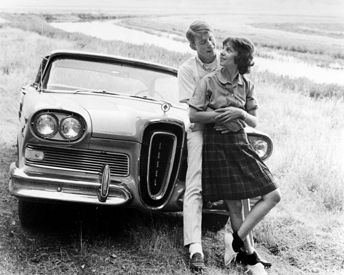 A black-and-white photo of a man holding a woman in front of a vintage car.