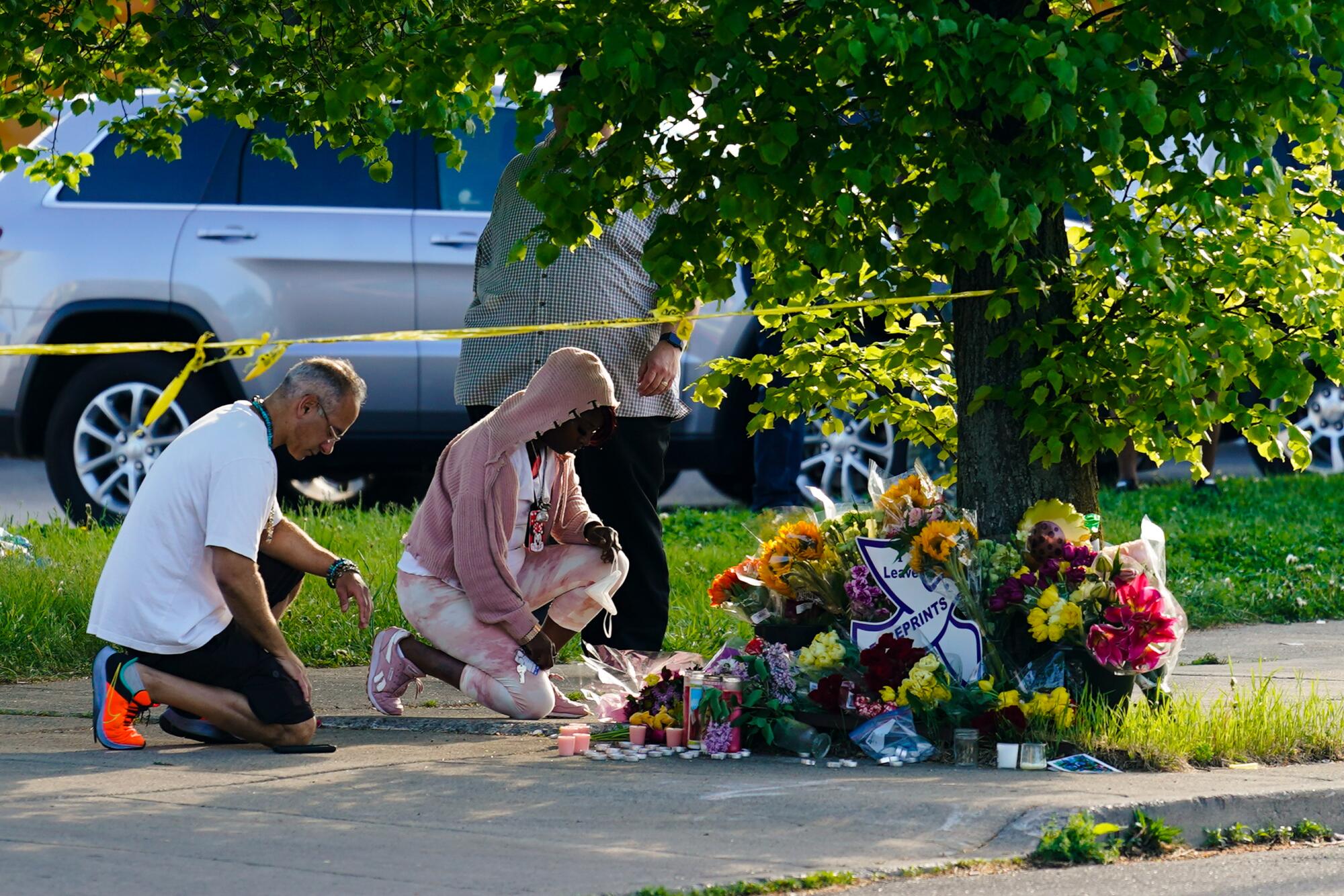 People kneel beside a sidewalk memorial of flowers near a tree and yellow police tape