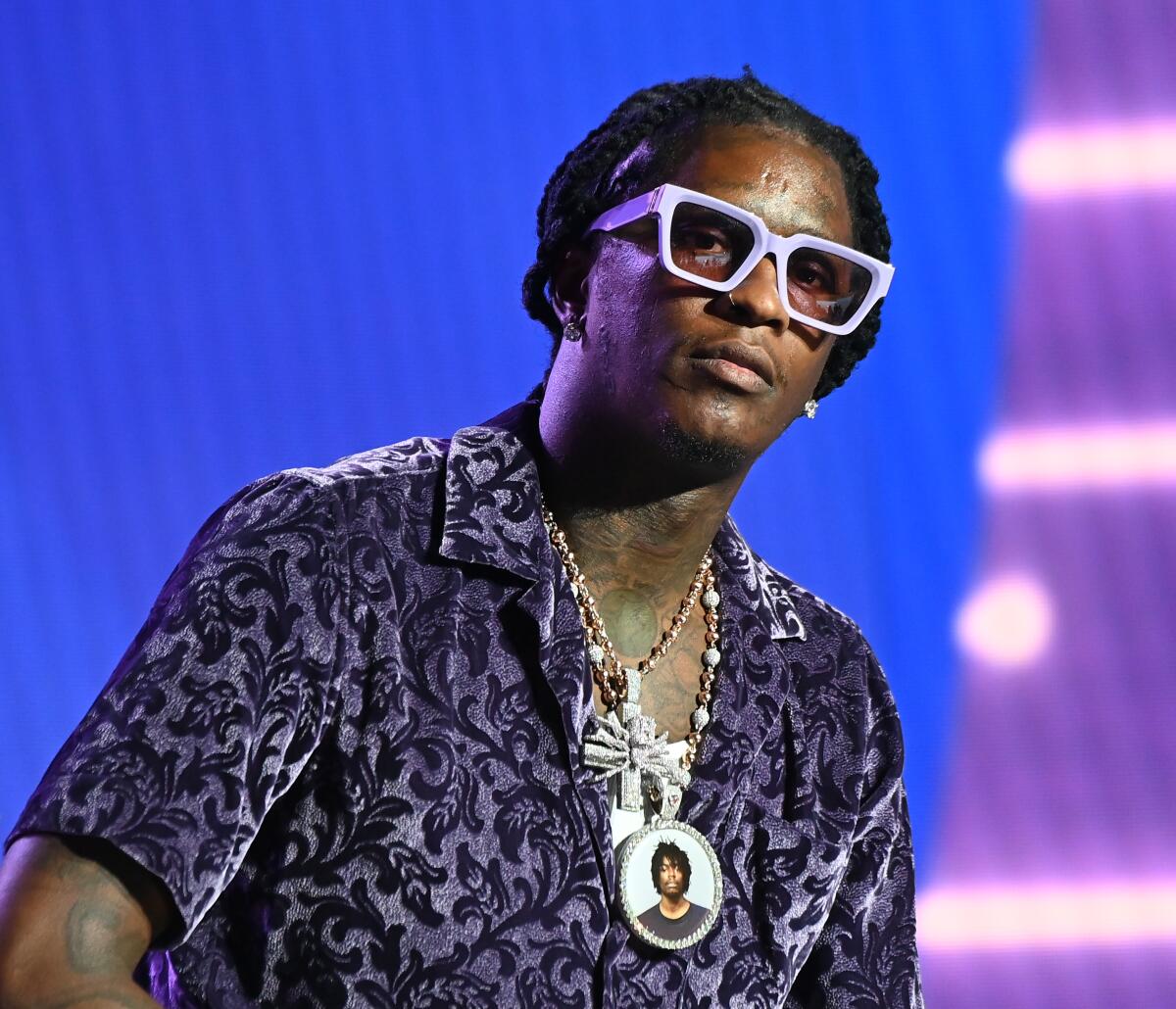 The rapper Young Thug wearing thick, white glasses, a dark patterned shirt and chains leaning forward and looking ahead