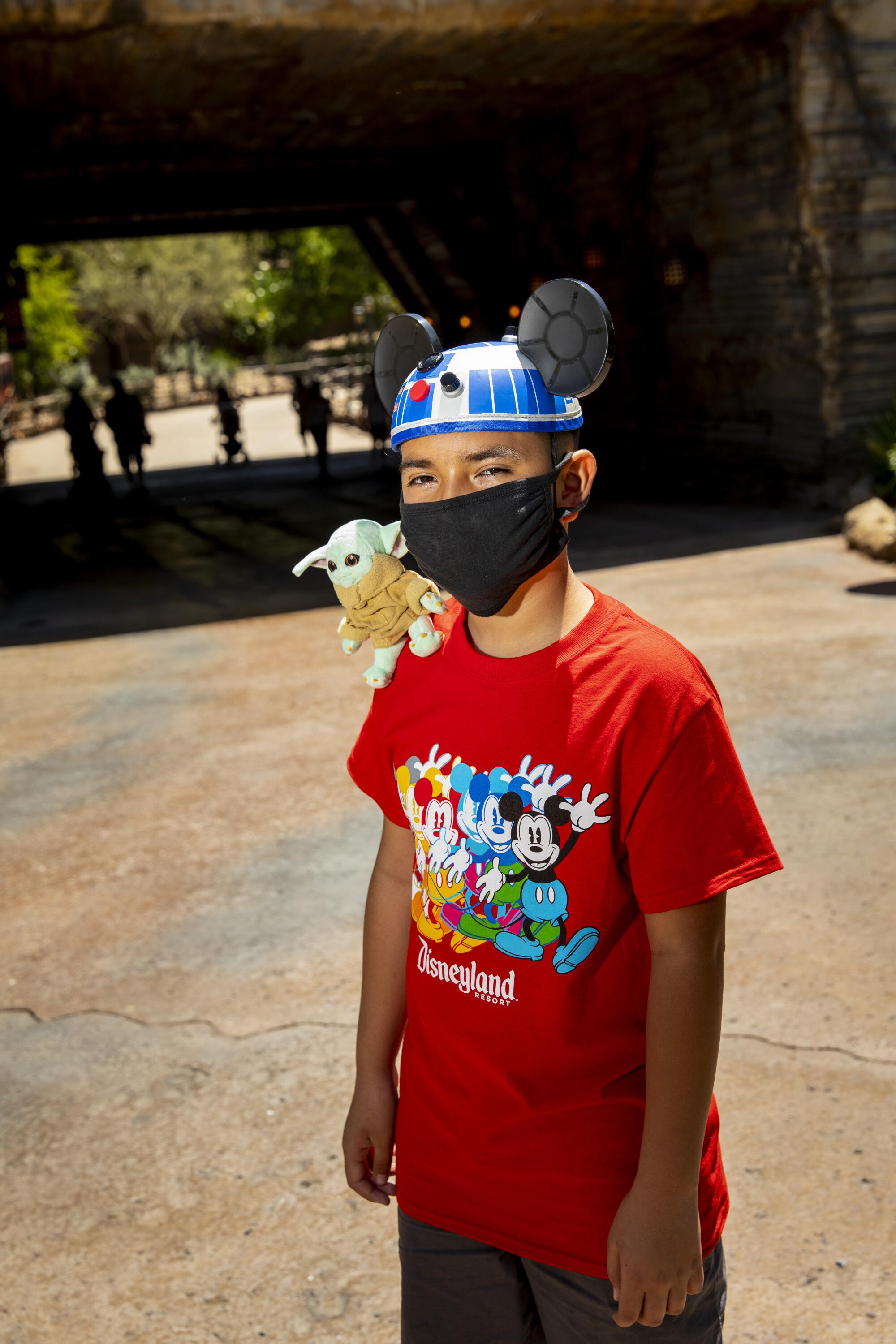 A boy in a Disneyland shirt and R2-D2 hat has a baby Yoda on his shoulder