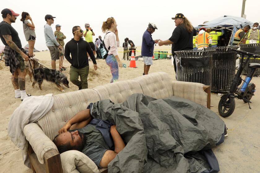 VENICE, CA - JULY 30, 2021 - - A homeless man sleeps on a couch while sprectators watch as sanitation crews clear a homeless encampment along Ocean Front Walk in Venice on July 30, 2021. The sanitation crew eventually woke the man and removed the couch. Over a dozen sanitation workers with the City of Los Angeles and contract workers with Beaches and Harbors clear homeless encampments and debris a few yards at a time on the beach near Thornton Avenue and Ocean Front Walk. The sweep began around 8 a.m. as members of St. Joseph Center and LAHSA tried to arrange housing for homeless who have been living on the beach. (Genaro Molina / Los Angeles Times)