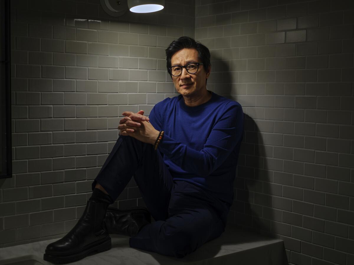 A man in glasses and a blue shirt leans against a corner wall as he sits on a bench with his hands on his knee.