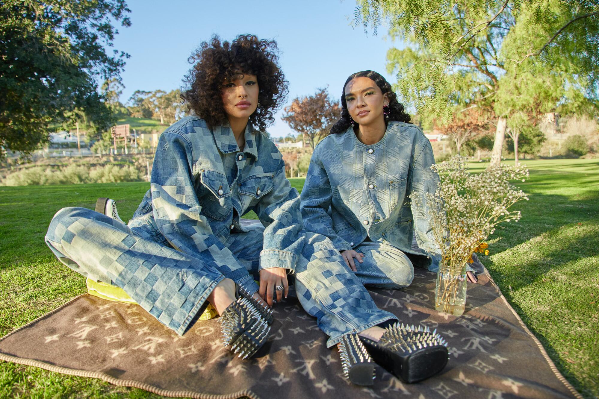 Two models wear Louis Vuitton denim ensembles on a picnic blanket in the park.
