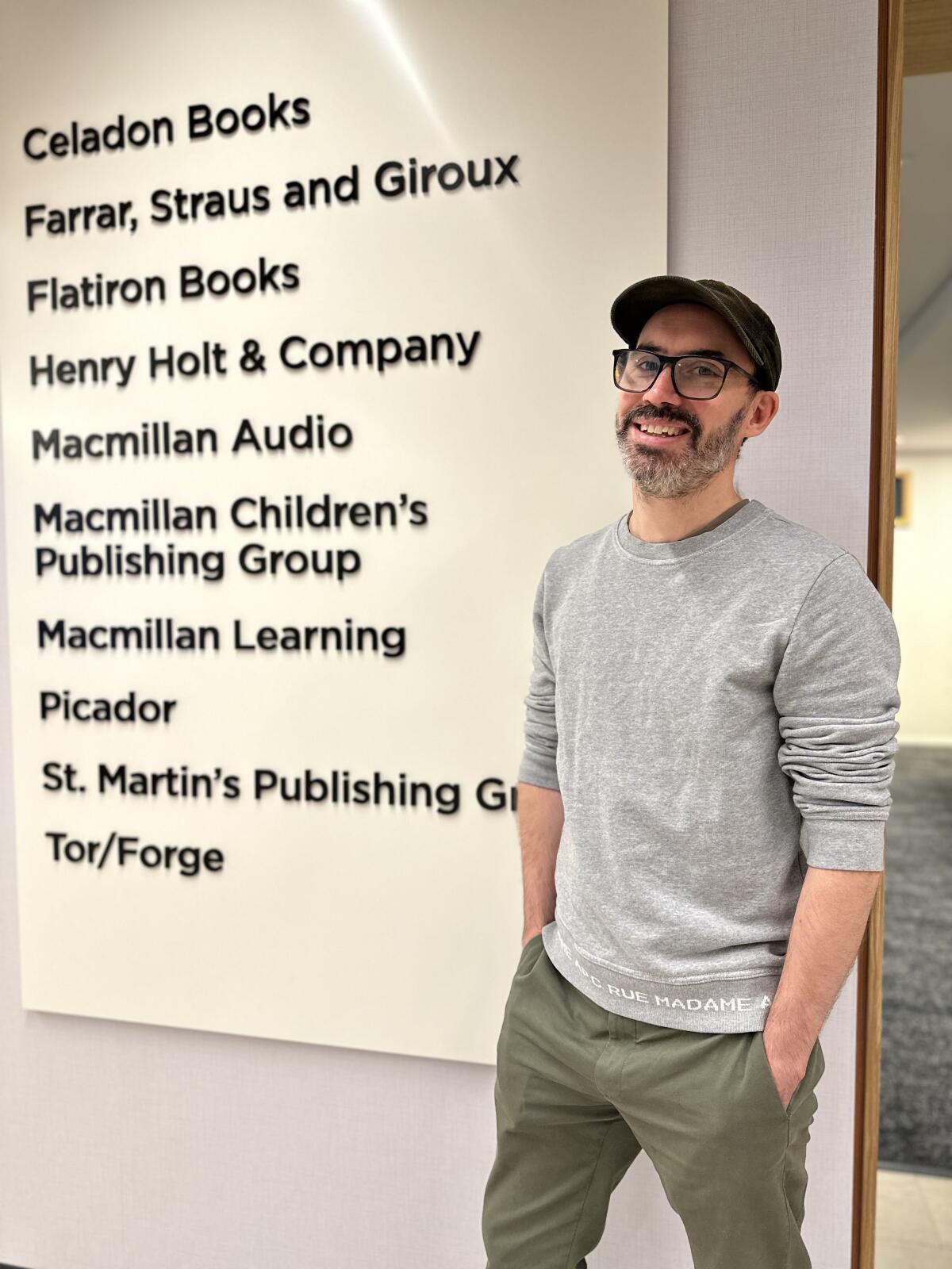 A man in a cap and grew sweater standing in front of a board listing the imprints of Macmillan Publishing.