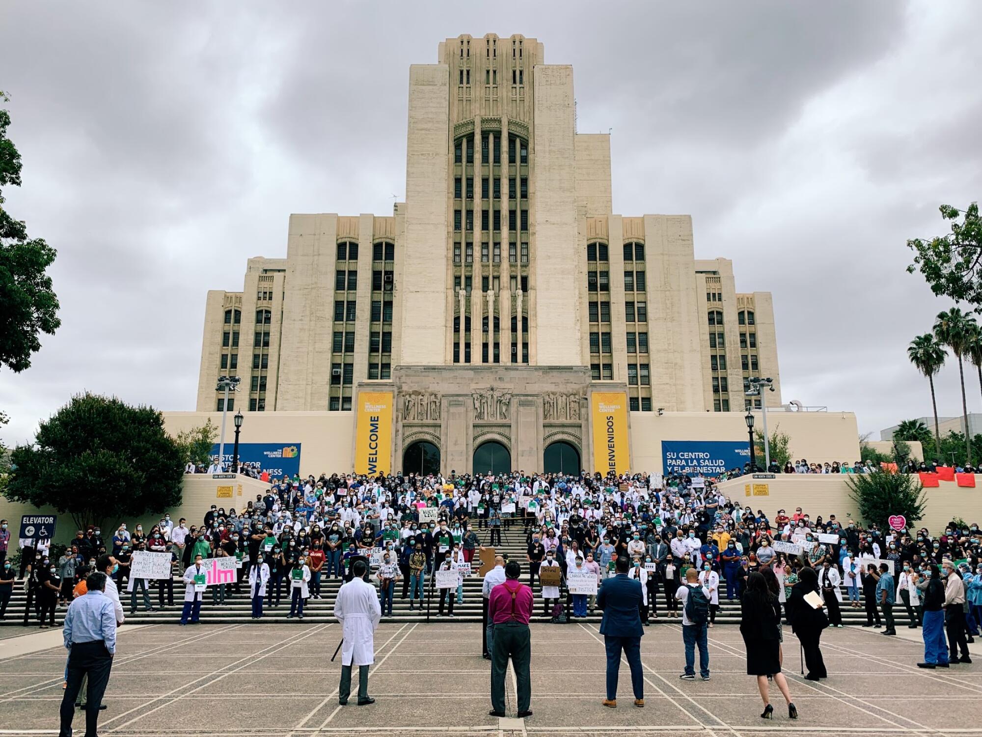 A large crowd of people with signs stand on the steps of a hospital building