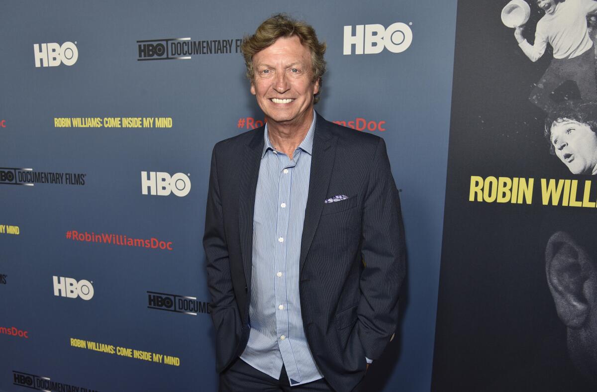 Nigel Lythgoe smiles and poses with his hands in pockets while clad in a dark blue suit jacket and a light blue dress shirt