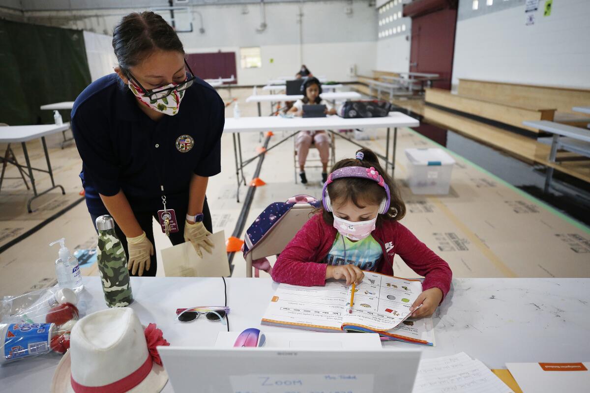 A first-grade girl does school work at a table as an adult aide looks on.