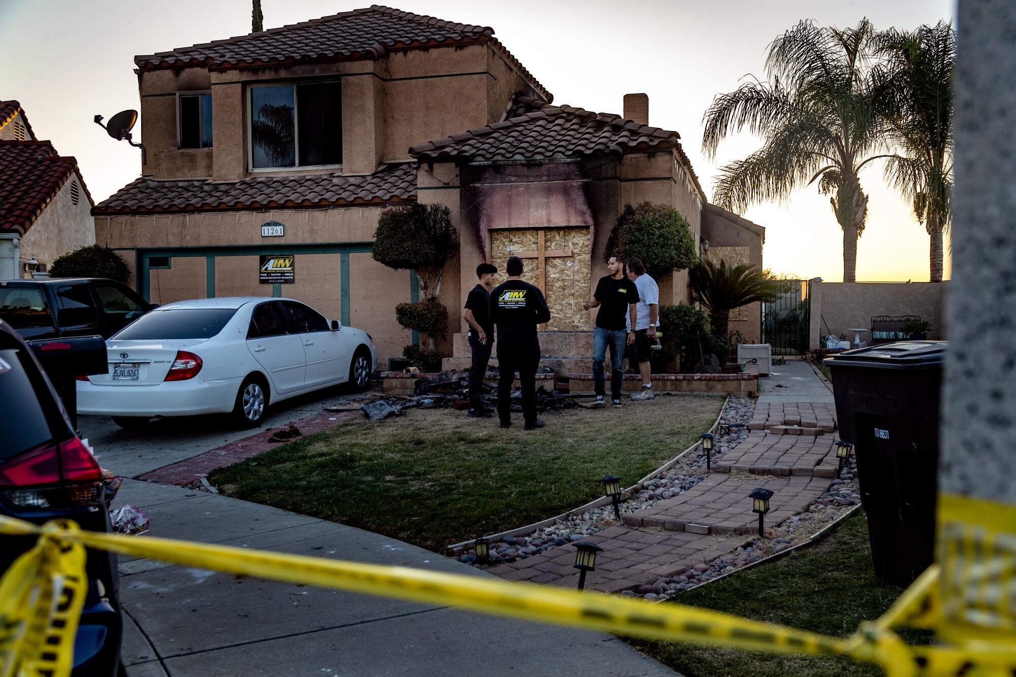 Four people stand on the front lawn of a burned home at dusk. Police crime tape cuts through the bottom of the frame.