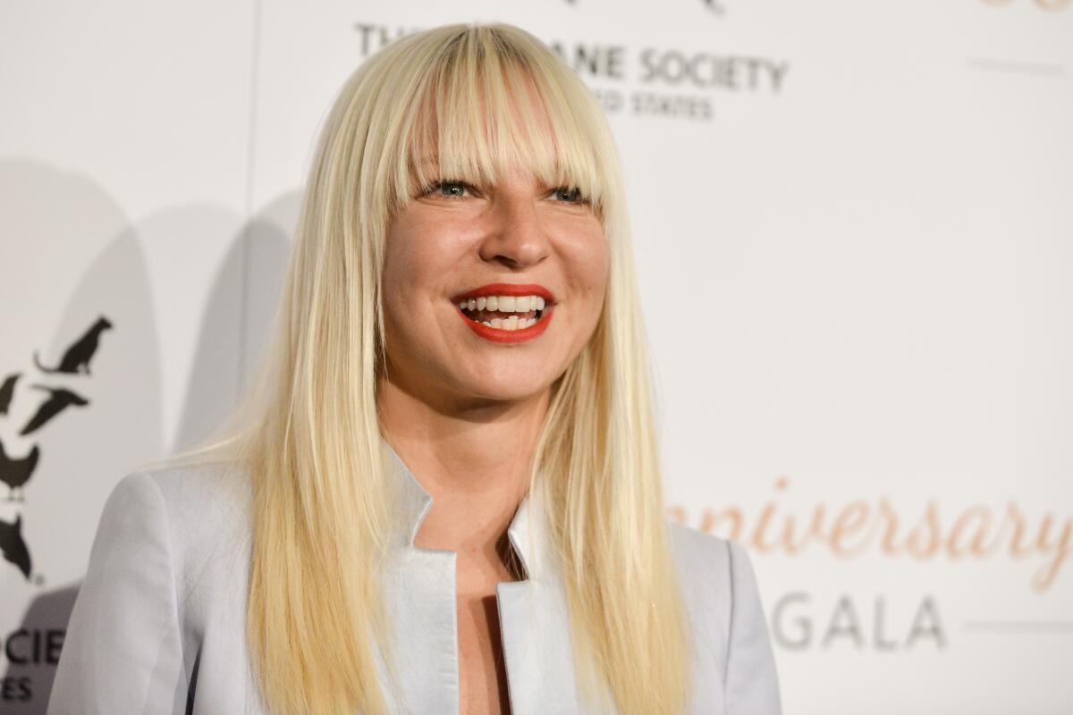 A smiling woman with long blond hair and bangs, wearing a light gray blazer 