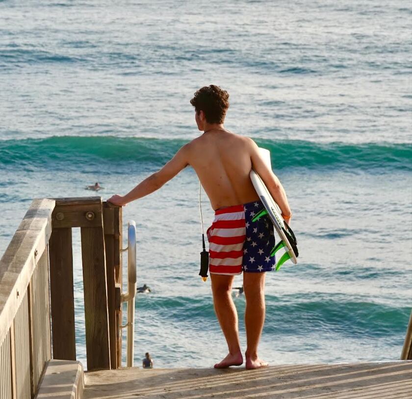 A man with a surfboard and American  flag swim trunks watches the waves