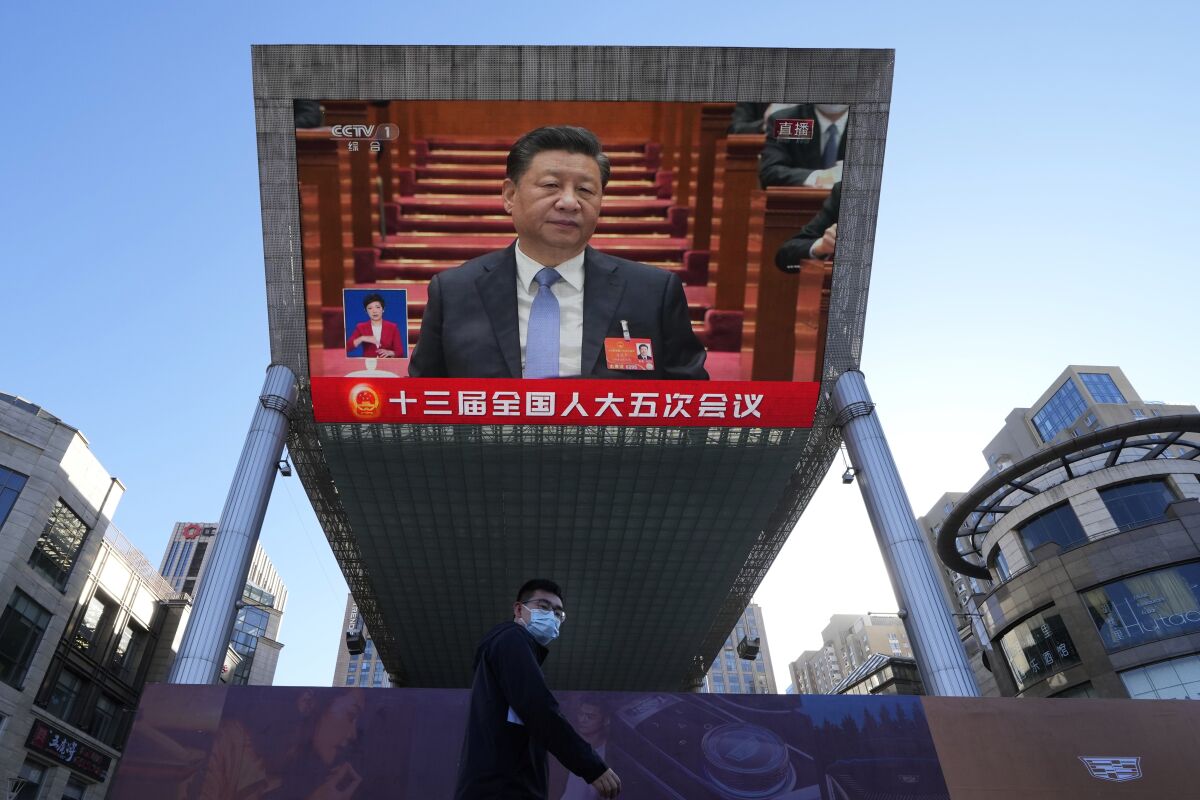 A masked pedestrian is seen under a large outdoor screen that shows a man with dark hair in a suit and light blue tie.