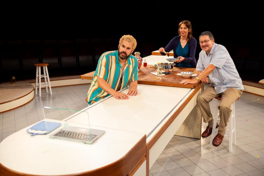 Three actors sit and stand around a kitchen counter in a theater.