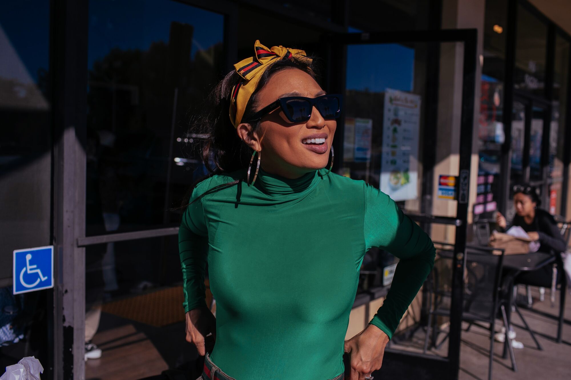A woman in sunglasses, a green top and a yellow head scarf smiles outside a restaurant. 
