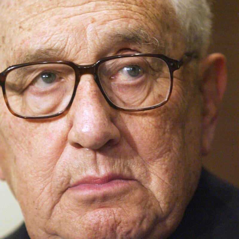 Henry Kissinger, with grey hair and glasses, looks toward his left