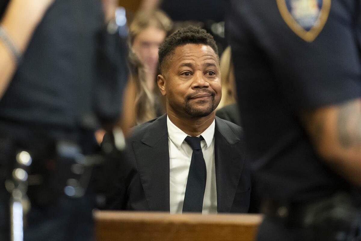 Cuba Gooding Jr. in a black suit and tie and white shirt sits in a courtroom.