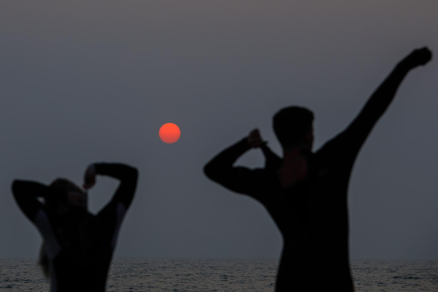 Two people get ready to surf as a hazy red sun sets off Hermosa Beach.