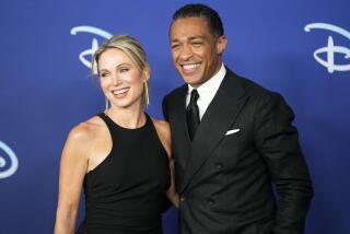 A blond woman in a black dress and a dark-haired man in a black suit smiling and posing for a photo together